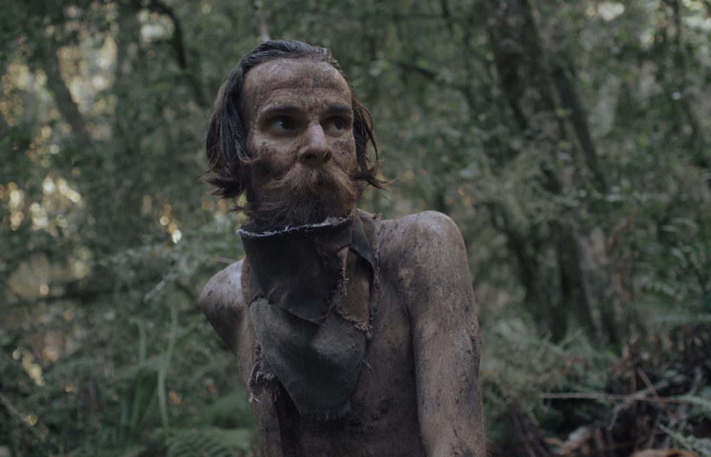 A skinny man covered in mud looks afraid or wary in a forest